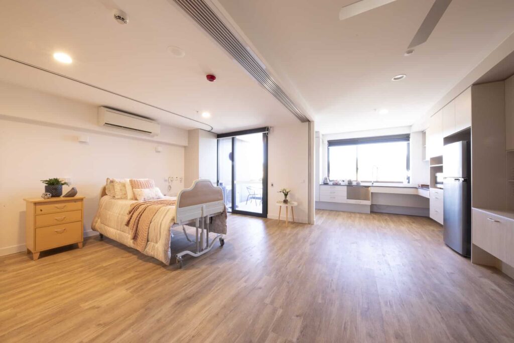 Bright, spacious hospital room with wood flooring, a single bed, bedside table with a plant, large windows, a balcony, built-in desk, storage cabinets, and modern lighting.