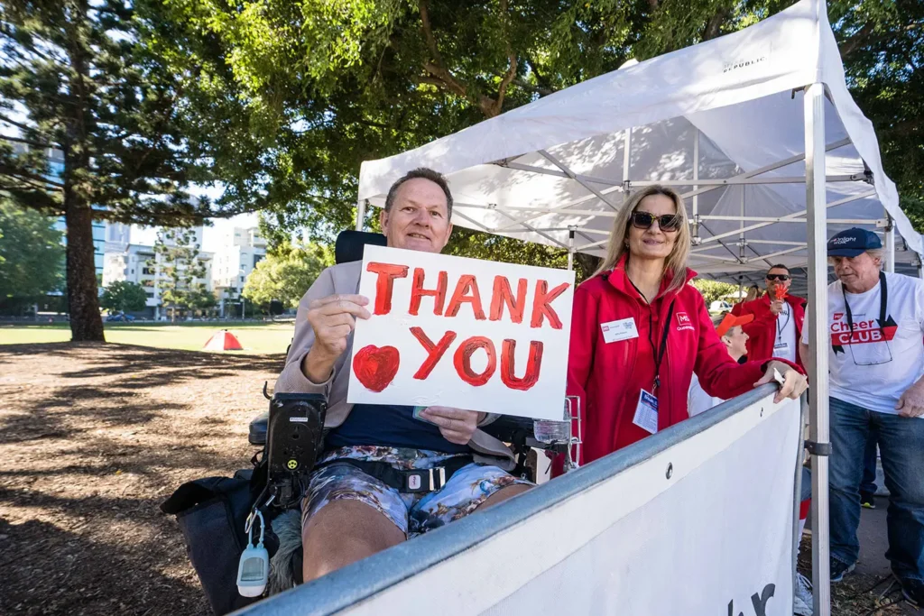 A person sitting in a wheelchair holds a sign that says "THANK YOU" with a red heart, while a woman in a red jacket stands beside him under a white tent at a park event.