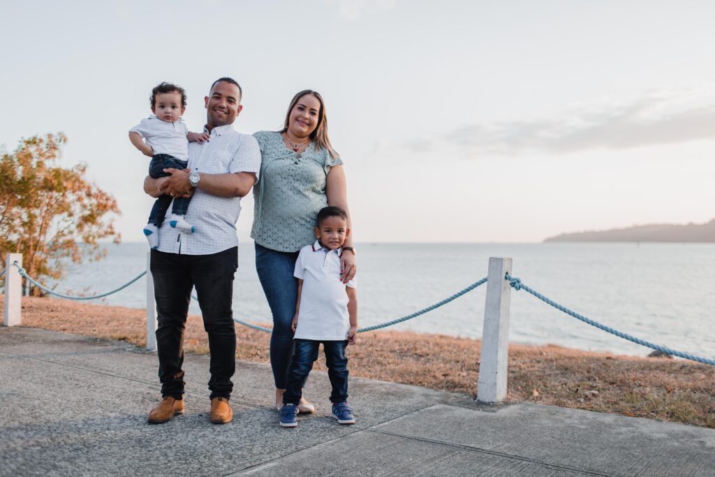 A family of four stands on a path by the water. The father holds a toddler while the mother stands next to him with an older child by her side. They all smile, dressed in casual clothes, with a scenic, calm waterfront in the background.