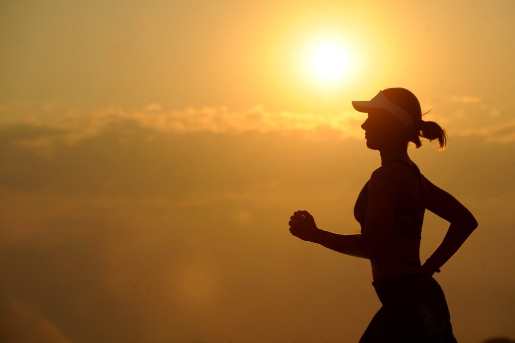 Silhouette of a person jogging outdoors at sunrise or sunset, with the sun shining brightly in the sky and soft clouds in the background.