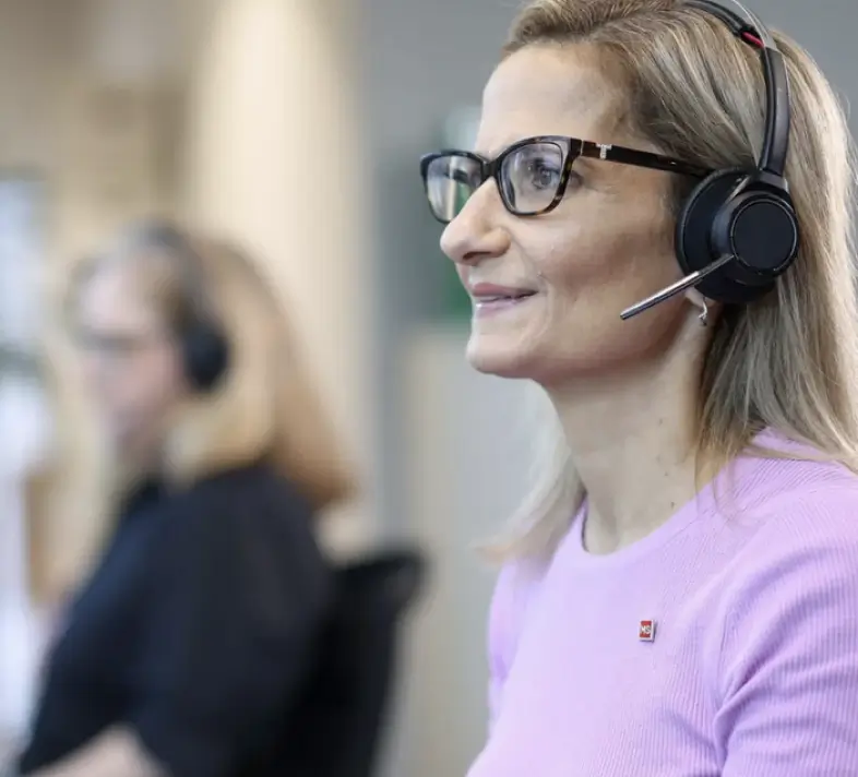 Woman in glasses and a headset, wearing a light purple top, sits at a desk and smiles while looking at a screen. Another person with a headset is blurred in the background.