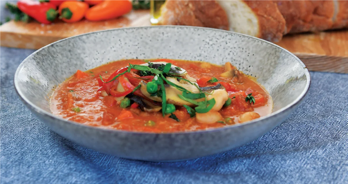 A bowl of vegetable stew with tomatoes, beans, and herbs, garnished with fresh green herbs, sits on a blue tablecloth. Sliced bread and colorful vegetables are visible in the background.