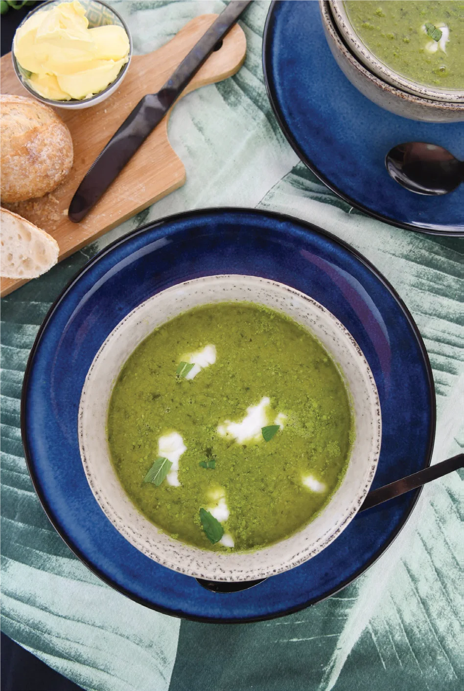A bowl of green soup garnished with cream and herbs sits on a blue plate, next to a spoon. Nearby are a cutting board with bread rolls, slices of bread, and a dish of butter.