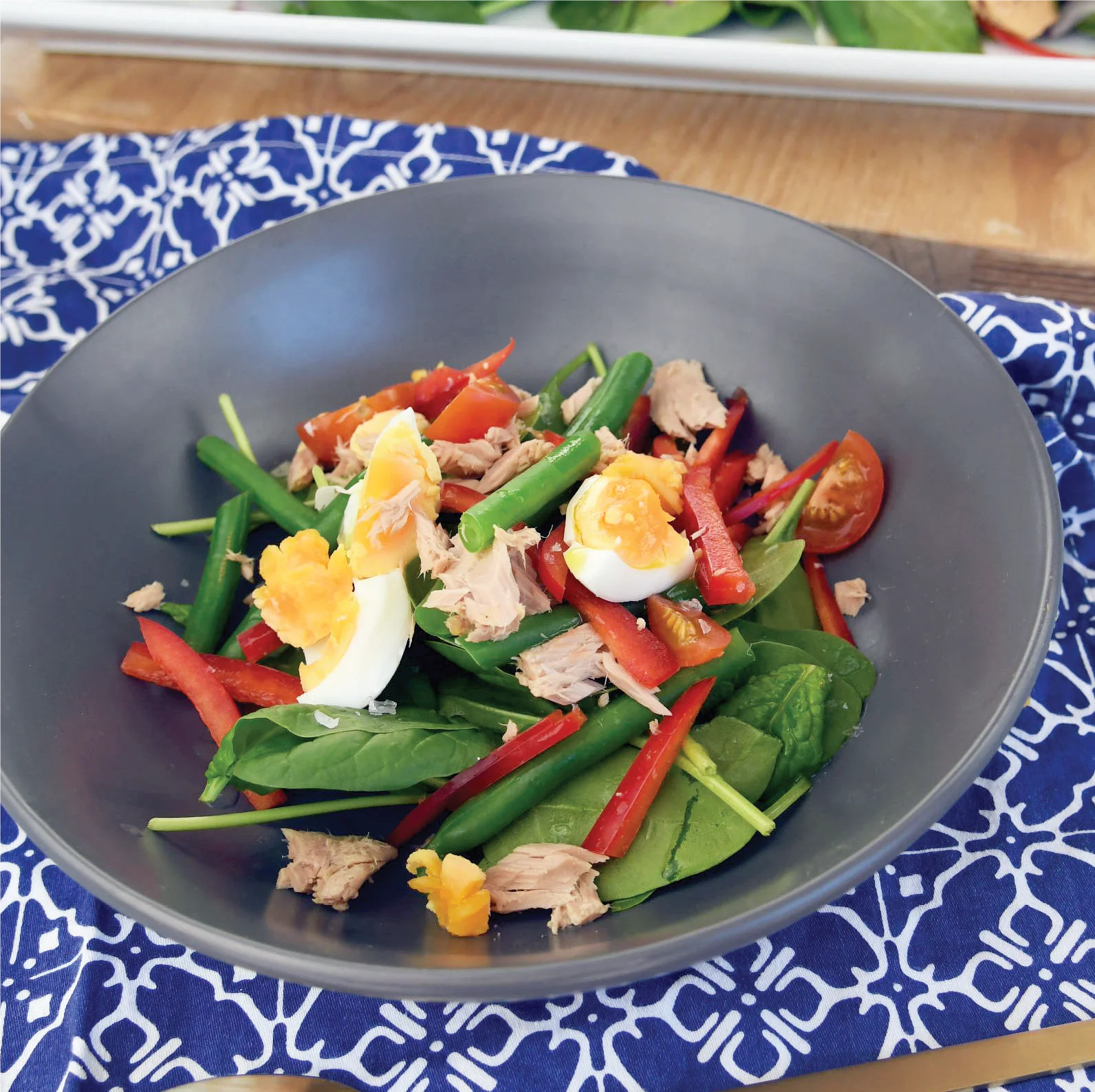 A gray bowl filled with salad containing spinach leaves, red bell pepper strips, green beans, cherry tomato halves, tuna chunks, and quartered hard-boiled eggs sits on a blue patterned cloth.