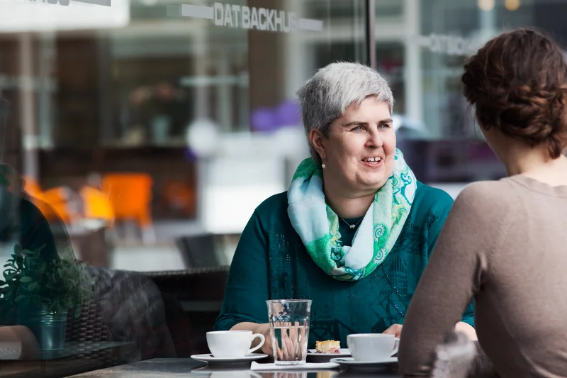 Two women are sitting at an outdoor café table, engaged in conversation. One woman with short gray hair and a green scarf is speaking, while the other listens. There are cups, glasses, and a plate on the table.