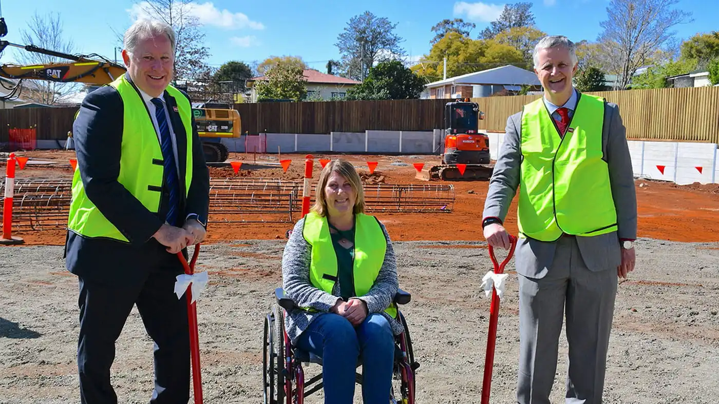 Three people wearing yellow safety vests, including one person in a wheelchair, hold shovels at a construction site with machinery and orange safety markers in the background.