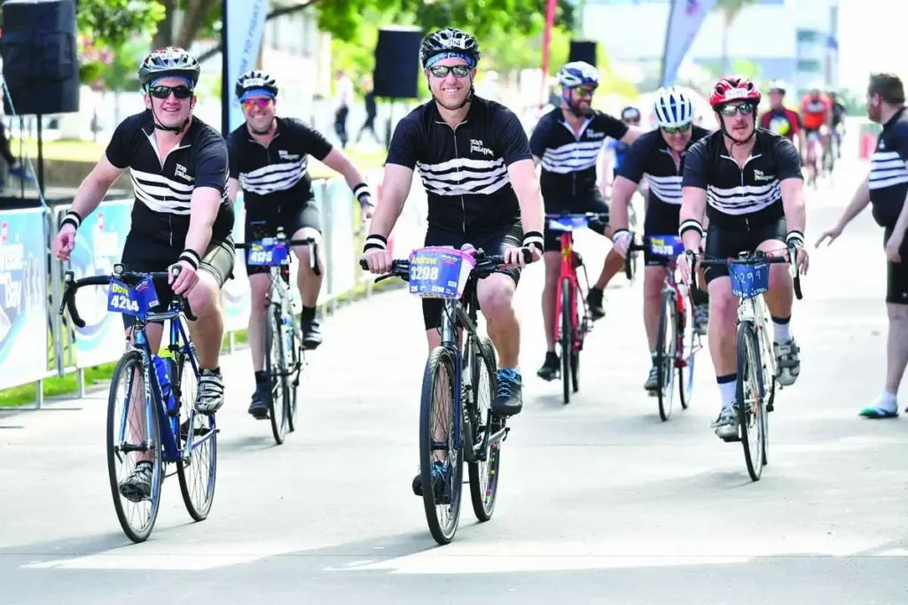 A group of six cyclists wearing matching black and white jerseys ride together during a cycling event on a sunny day, with event banners and other participants visible in the background.