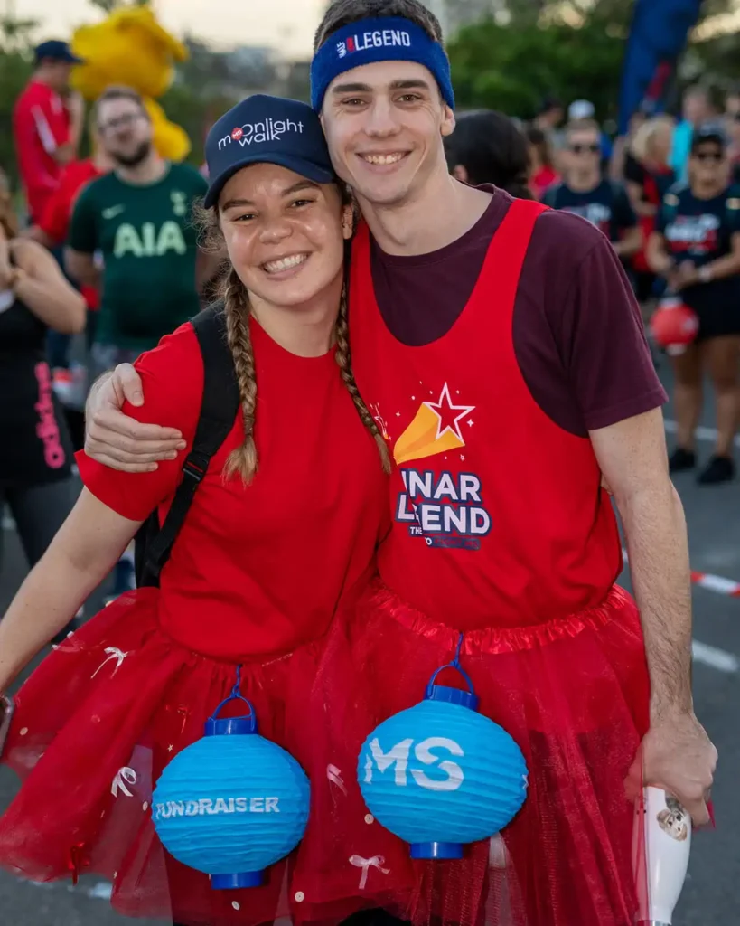 Two people wearing red shirts, red tutus, and blue paper lanterns labeled "Fundraiser" and "MS" at a festive outdoor event, smiling with arms around each other. Other participants and colorful outfits in the background.