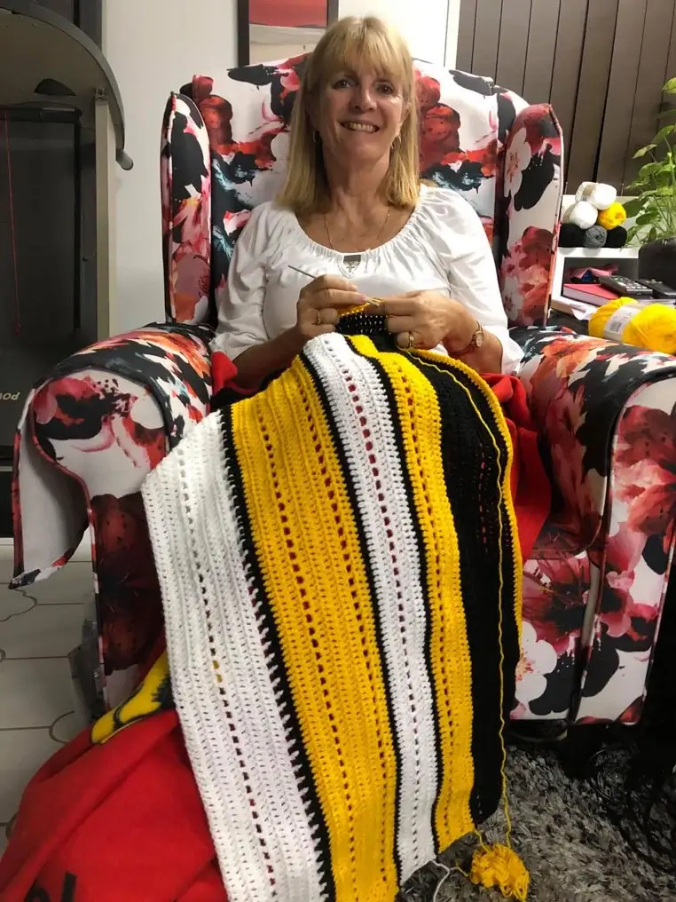 A smiling woman sits in a floral armchair, knitting a striped blanket in yellow, black, and white yarn. Balls of yarn rest beside her, and a red blanket is draped over her lap.