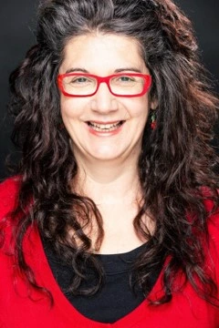 A woman with long, curly dark hair smiles at the camera. She is wearing red glasses, a red top over a black shirt, and has a dark background behind her.