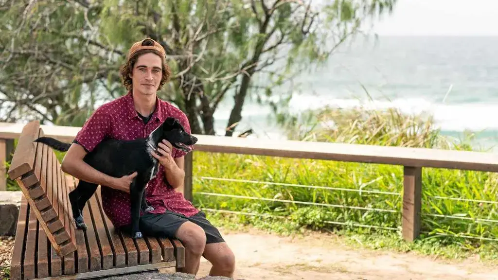 A young man in a red patterned shirt and baseball cap sits on a wooden bench by the beach, smiling and holding a black dog on his lap, with greenery and ocean waves in the background.
