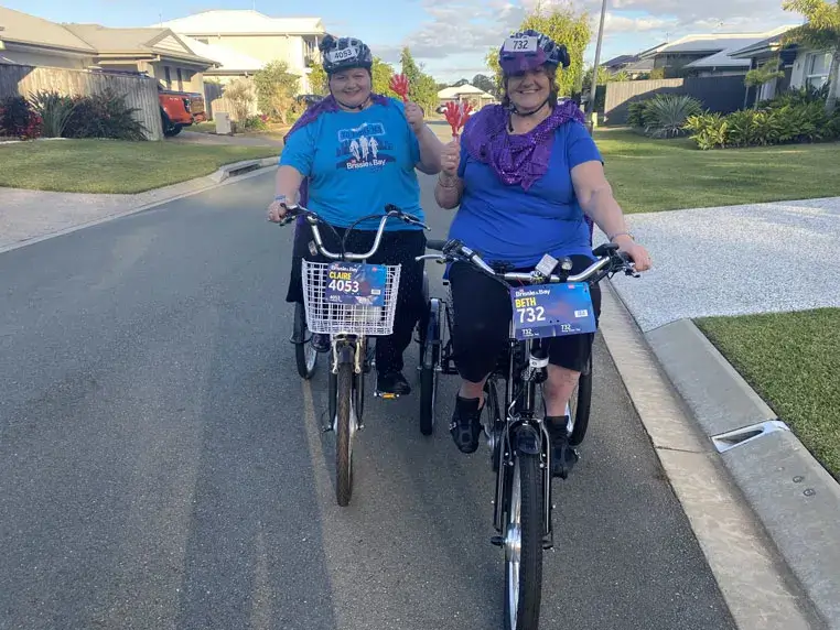 Two women wearing helmets and blue shirts smile as they ride bicycles down a suburban street. One woman waves at the camera. Both bikes have number plates attached to the front. Houses and greenery line the road.