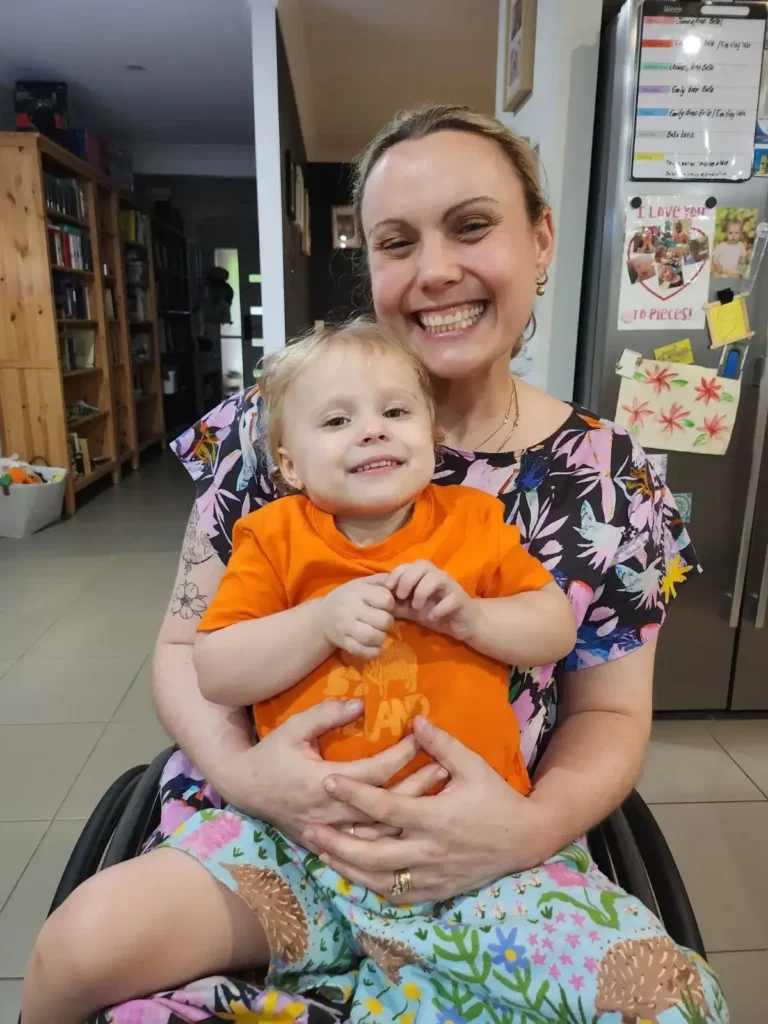 A smiling woman in a colorful dress sits in a wheelchair, holding a young child in an orange shirt. They are in a cozy kitchen with drawings and notes on the fridge in the background. Both look happy and relaxed.