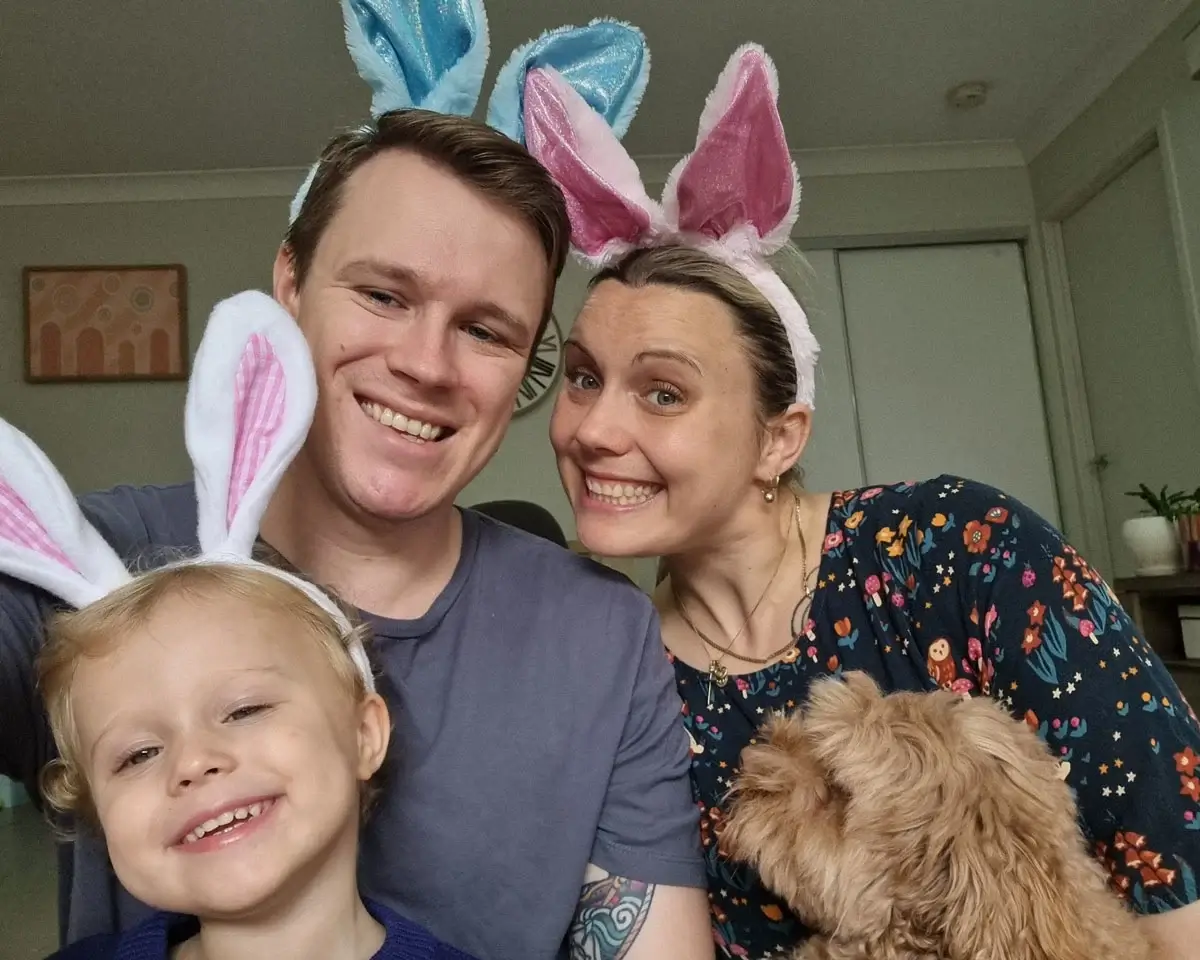 A smiling family of three, all wearing bunny ears headbands, poses for a selfie indoors with a small, fluffy brown dog in front of them. The background shows a light-colored room with a few decorations.