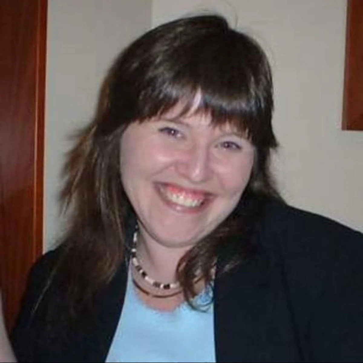A woman with straight brown hair and bangs smiles broadly while wearing a black blazer, light blue top, and a beaded necklace, standing indoors against a light-colored wall.
