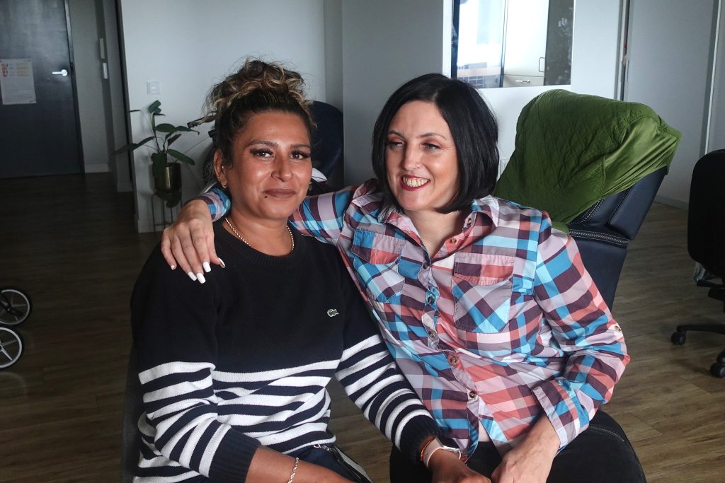 Two women sit closely together indoors, smiling at the camera. One wears a black and white striped sweater; the other wears a colorful plaid shirt. They appear relaxed and happy, with one woman's arm around the other's shoulder.