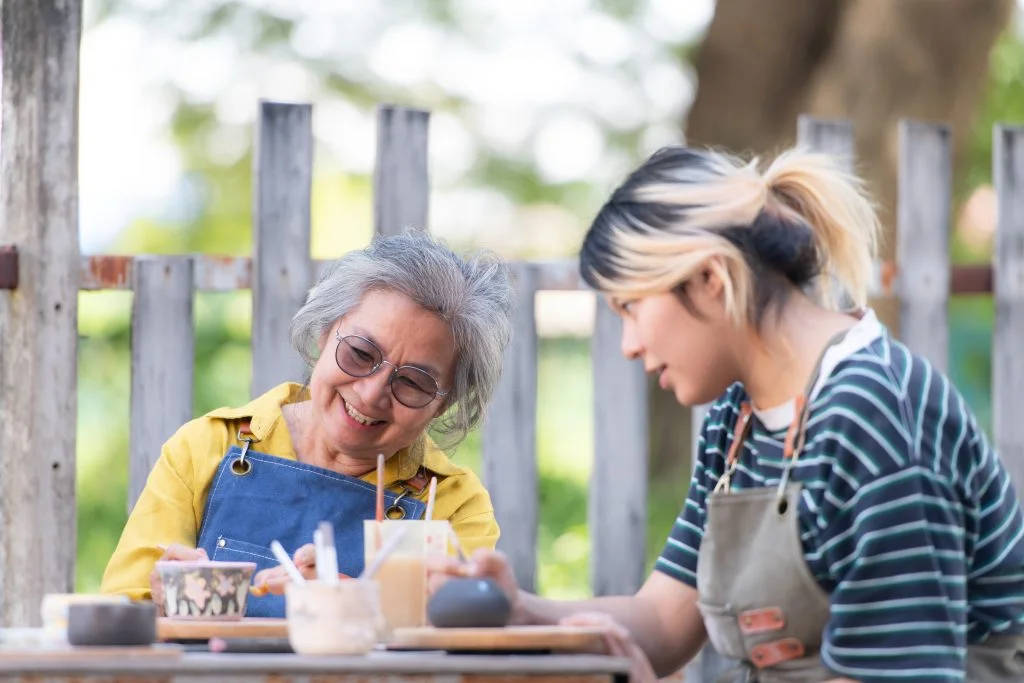 Two women, one older and one younger, sit outside at a table working on pottery and painting ceramics together. They both wear aprons and appear happy and focused on their creative activity.