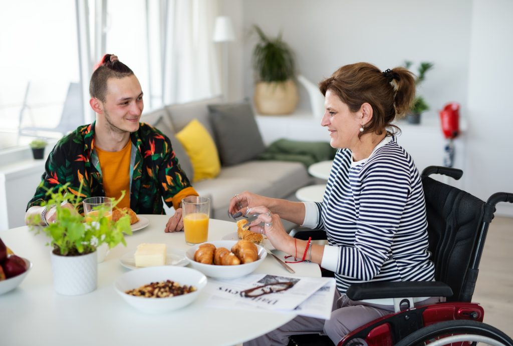 A woman in a wheelchair and a young man sit at a table with pastries, orange juice, and coffee, smiling and talking in a bright, cozy living room.