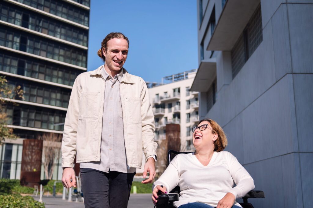 A man walks beside a woman in a wheelchair outdoors on a sunny day. Both are smiling and enjoying each other's company, with modern buildings in the background.