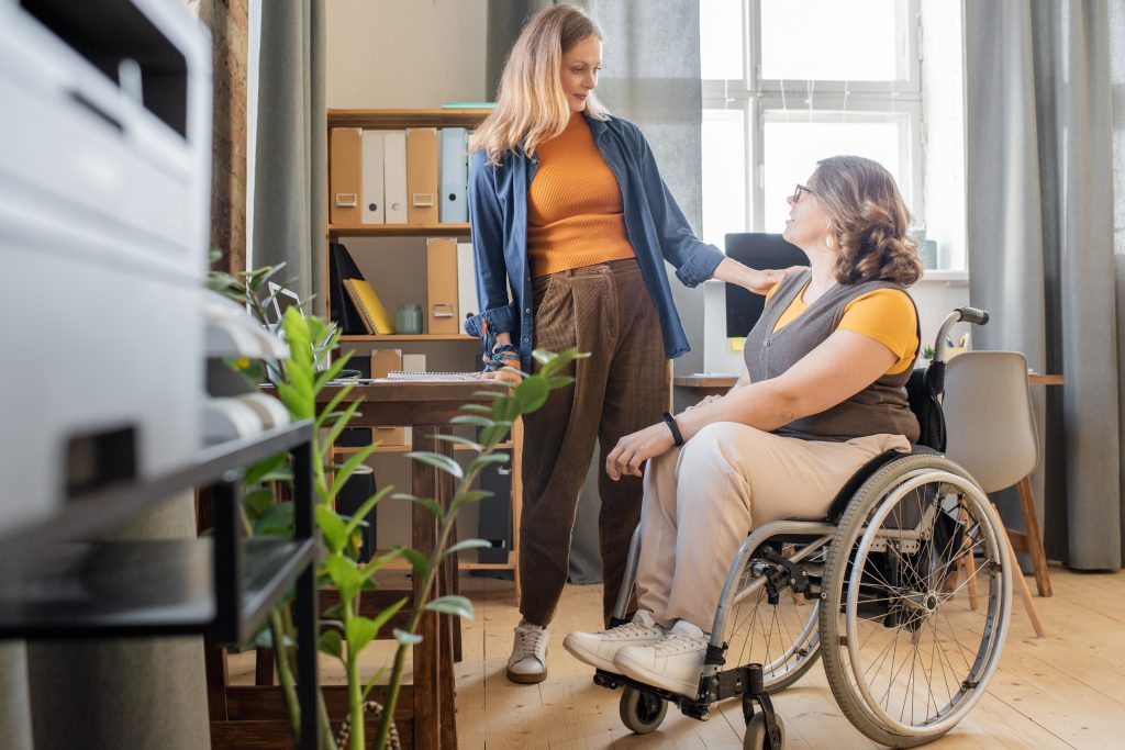 Two women in an office, one standing and the other sitting in a wheelchair. They are smiling and talking, with one woman resting her hand on the other's shoulder. There are shelves, a desk, and plants nearby.