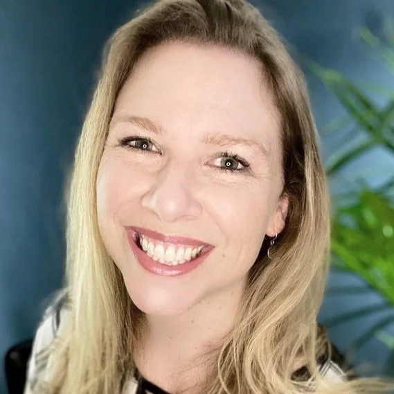 A woman with long blonde hair smiles at the camera. She is wearing a black-and-white top and standing in front of a blue background with a green plant visible on the right.