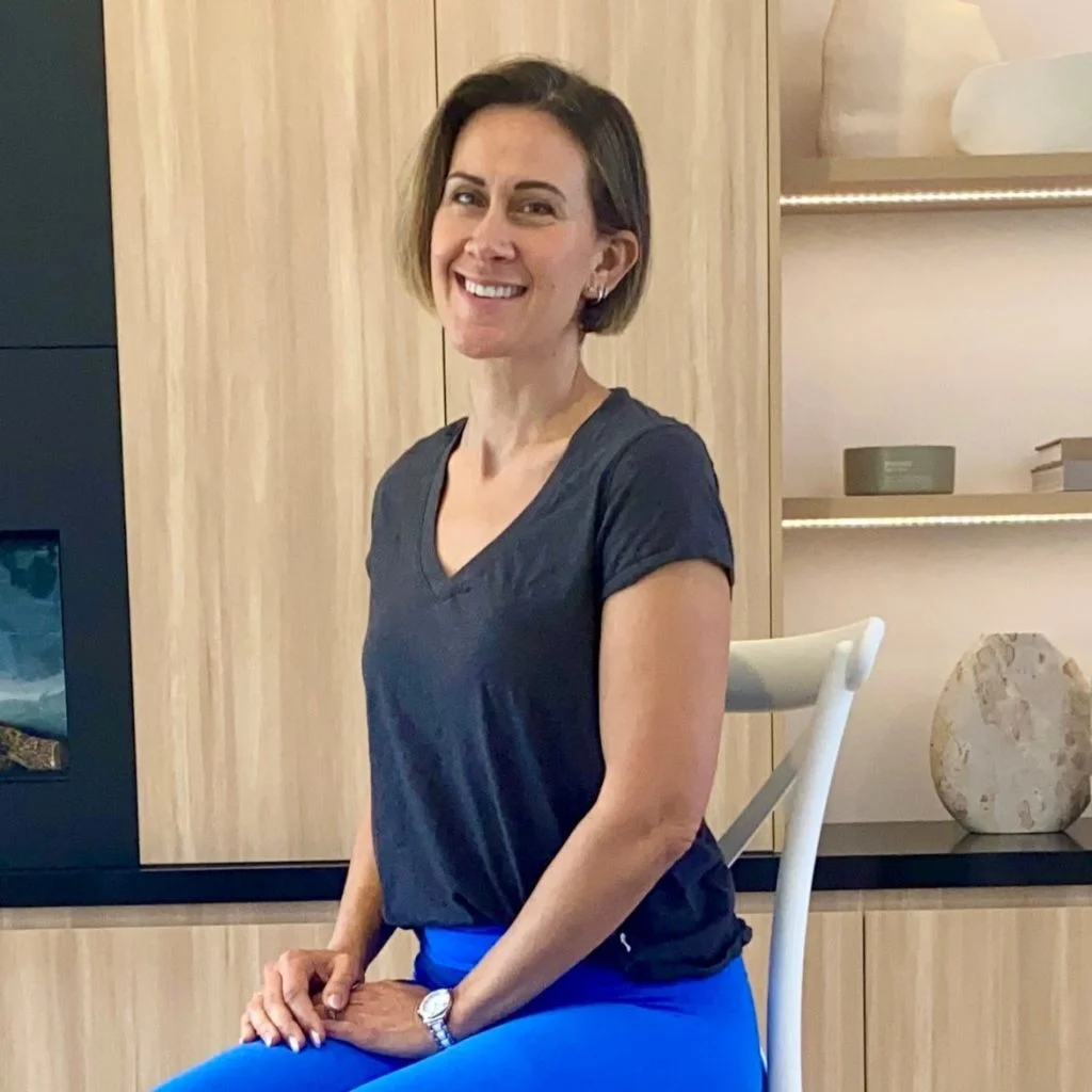 A smiling woman with short brown hair sits sideways on a white chair. She wears a dark gray V-neck shirt and bright blue pants. Behind her are wooden cabinets, a stone decoration, and books on a shelf.