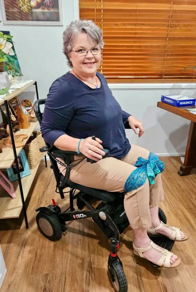 Smiling older woman with glasses and gray hair sits in a power wheelchair indoors. She wears a navy top, tan pants with a blue scarf tied around one knee, and sandals, with wooden shelves and a window in the background.