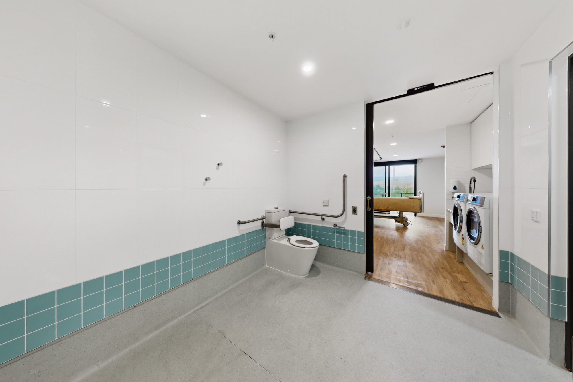 Accessible bathroom with a toilet and grab rails, leading into a room with laundry machines and a hospital bed.