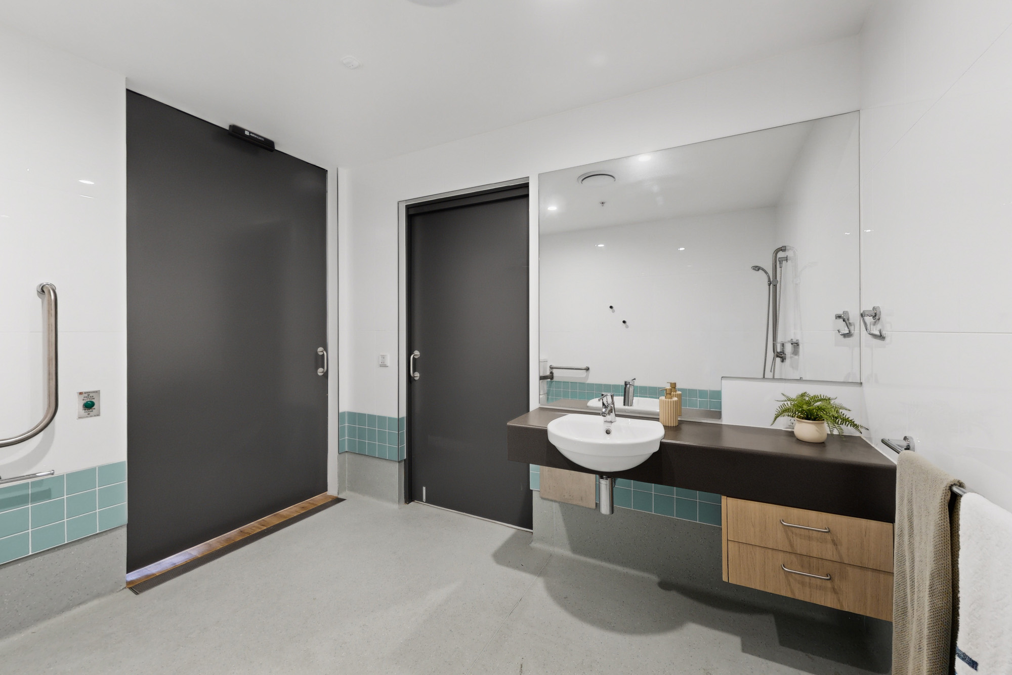 Accessible bathroom featuring a wall-mounted sink with a large mirror, dark grey doors, a grab bar, and light blue tiled accents.