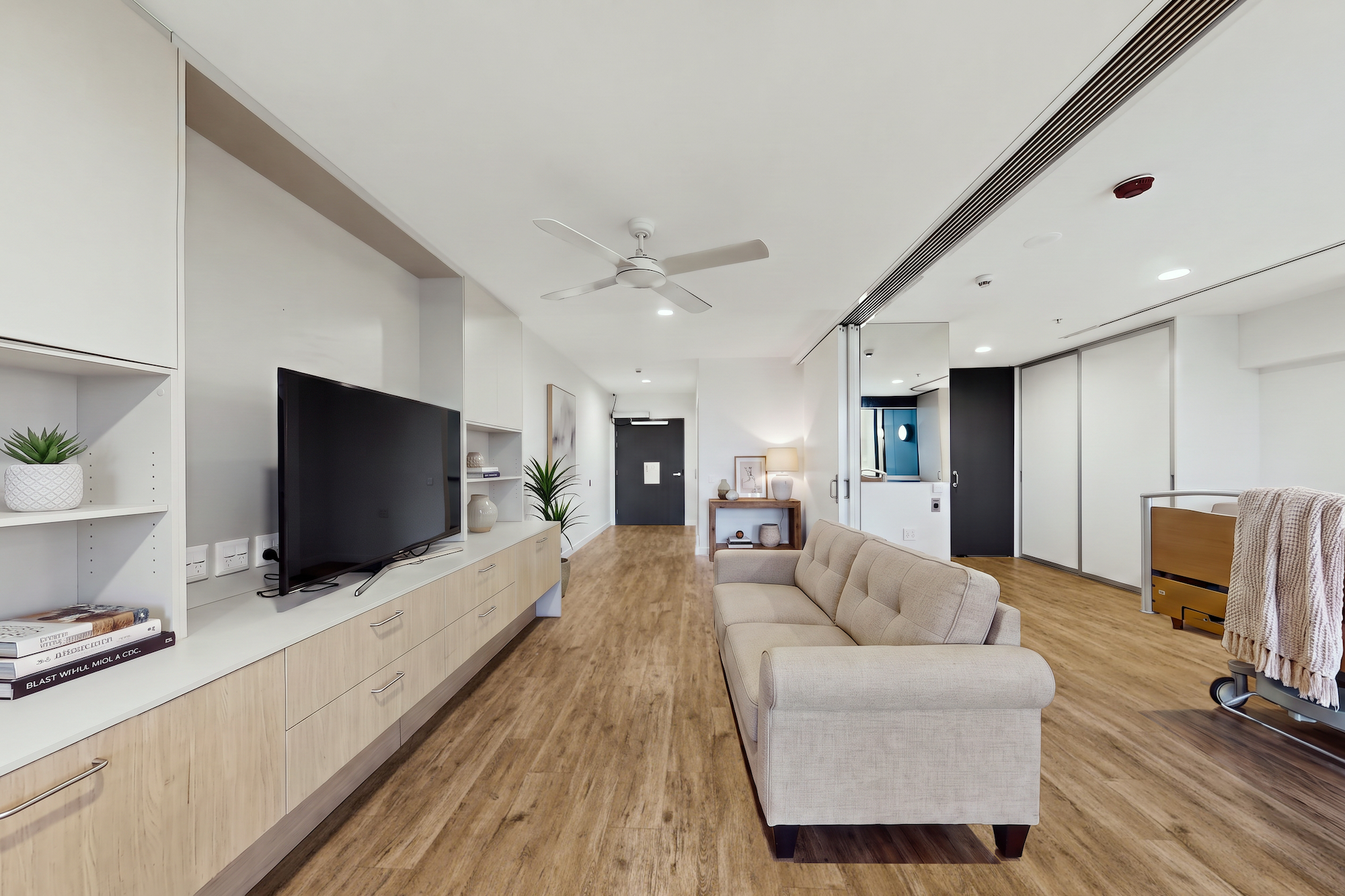 A modern living room with a neutral-colored sofa, a wall-mounted television on built-in shelving, and light wood flooring.