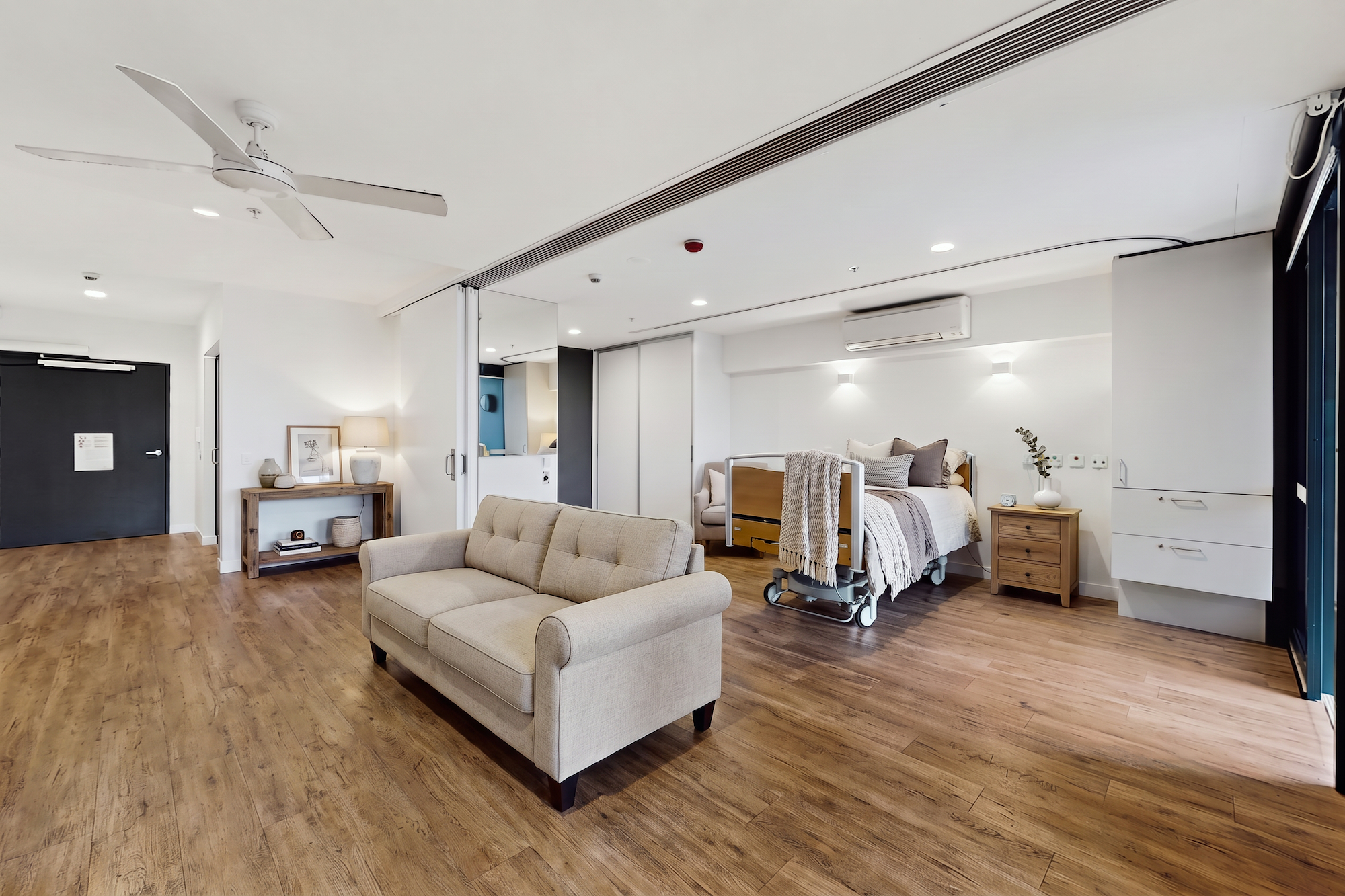 A spacious residential-style bedroom featuring a light-colored sofa in the foreground and a hospital-style bed with neutral bedding in the background.