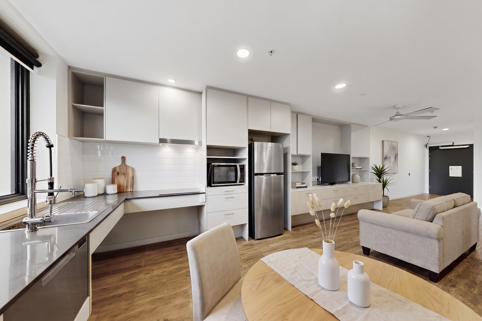 A modern apartment with an open-plan kitchen featuring stainless steel appliances, light gray cabinetry, and a dining area with a round wooden table, flowing into a living room with a beige sofa.