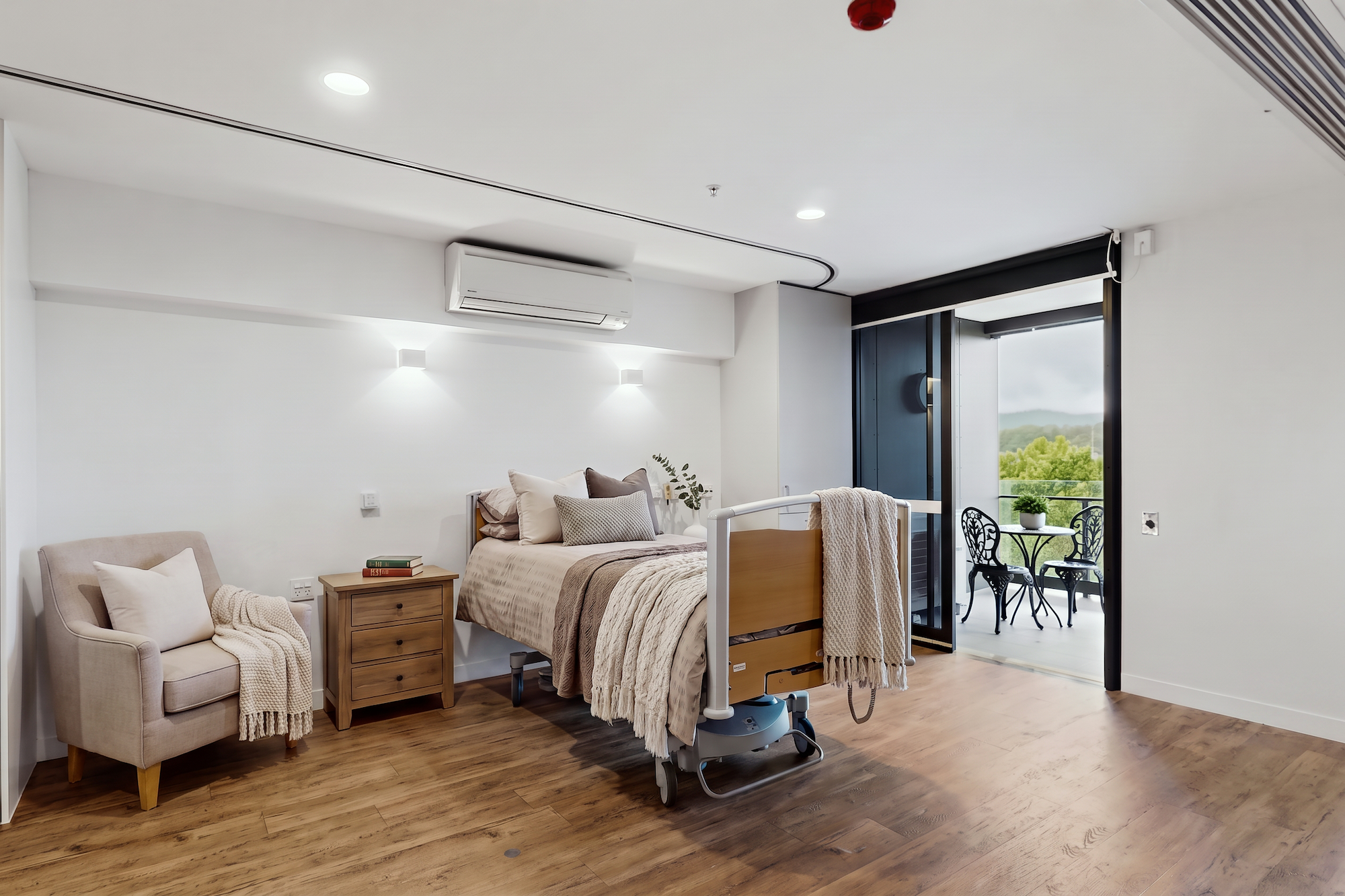 A modern hospital bedroom with a patient bed, a bedside table with books, an armchair, and a glass door opening to a balcony with bistro seating.
