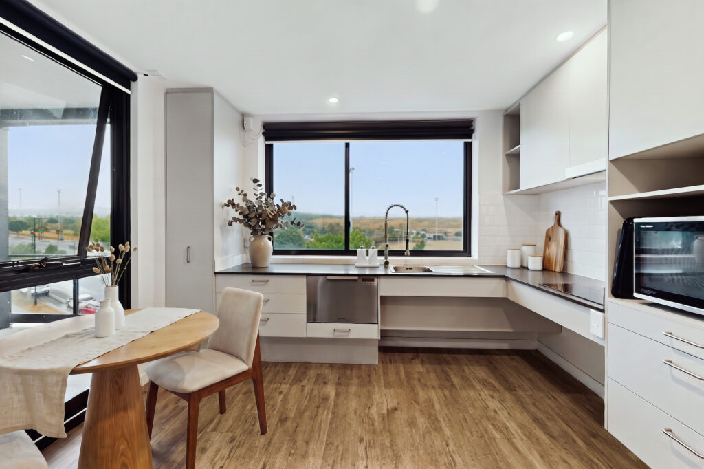 A kitchen with white cabinets, wood flooring, and a dining table, featuring a window overlooking a grassy field.