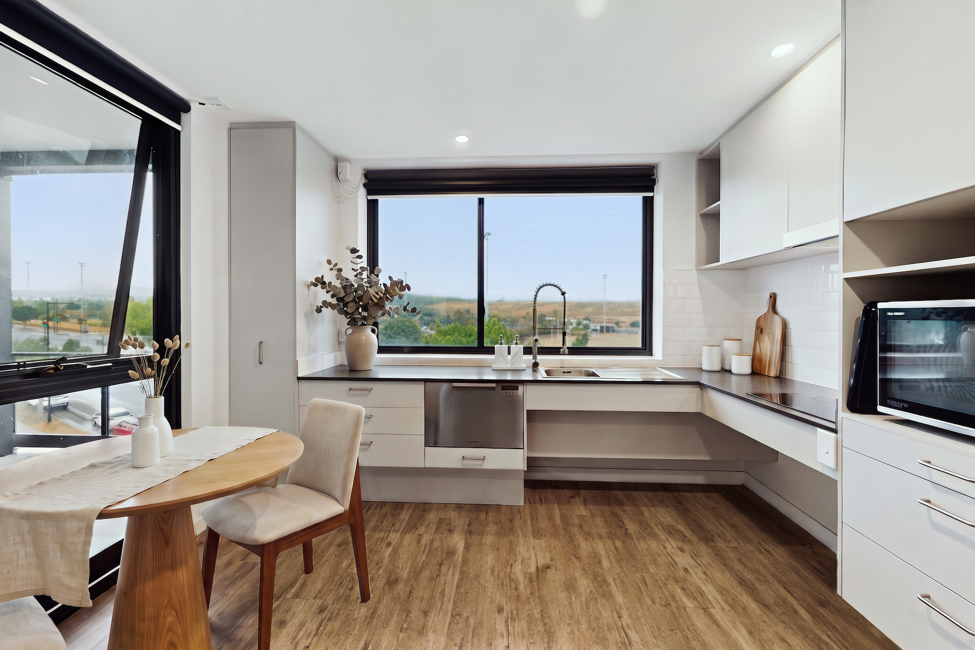 A kitchen with white cabinets, wood flooring, and a dining table, featuring a window overlooking a grassy field.