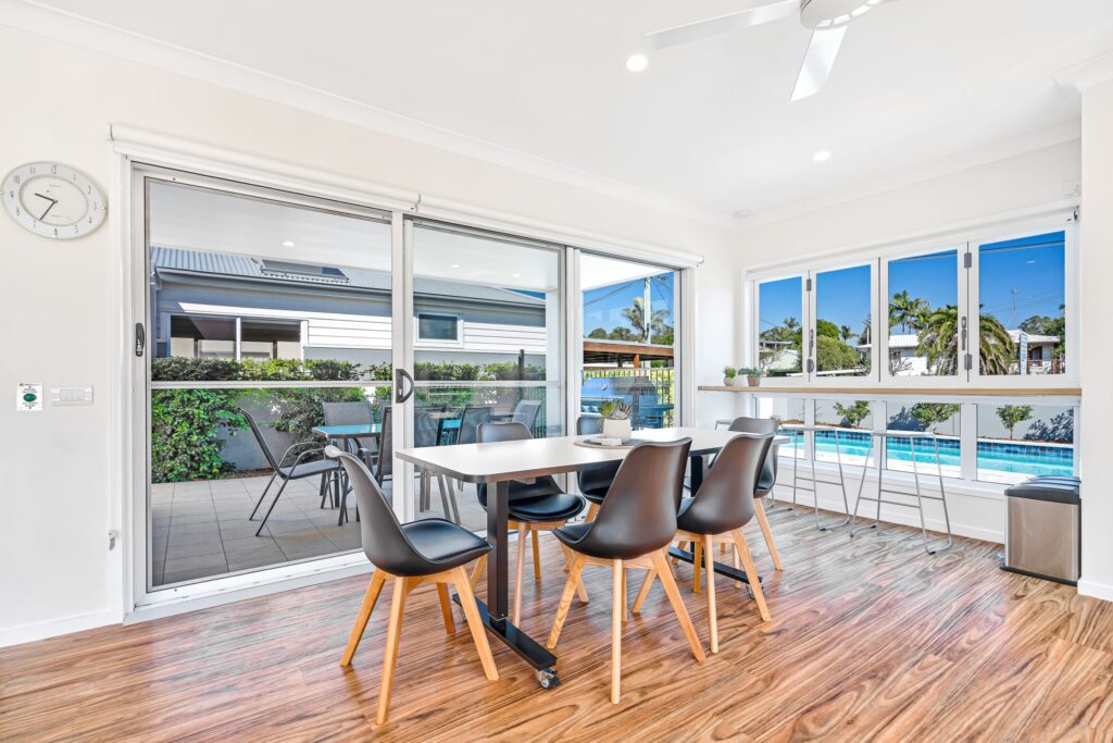 Modern dining area with black chairs and a white table, large sliding glass doors opening to an outdoor patio, and windows overlooking a swimming pool and garden. The room has wood flooring and is bright with natural light.