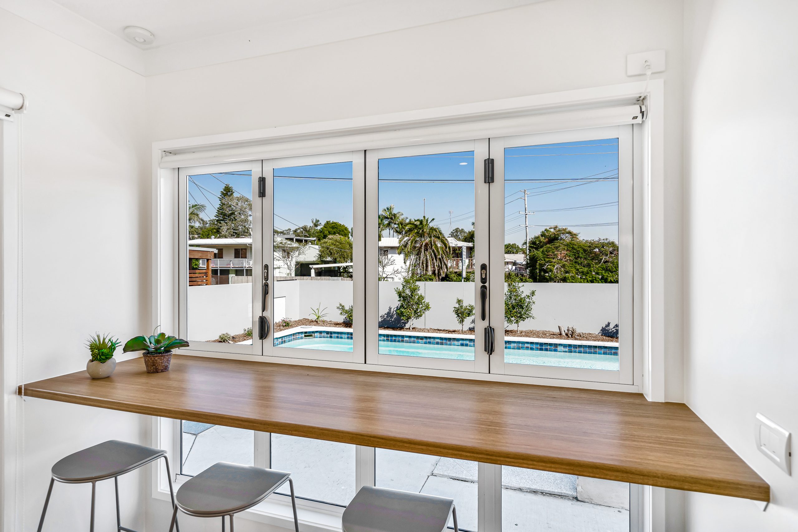 Modern kitchen bar with wooden countertop and two stools beside a wide window, overlooking a backyard with a swimming pool, palm trees, and neighboring houses under a clear blue sky.