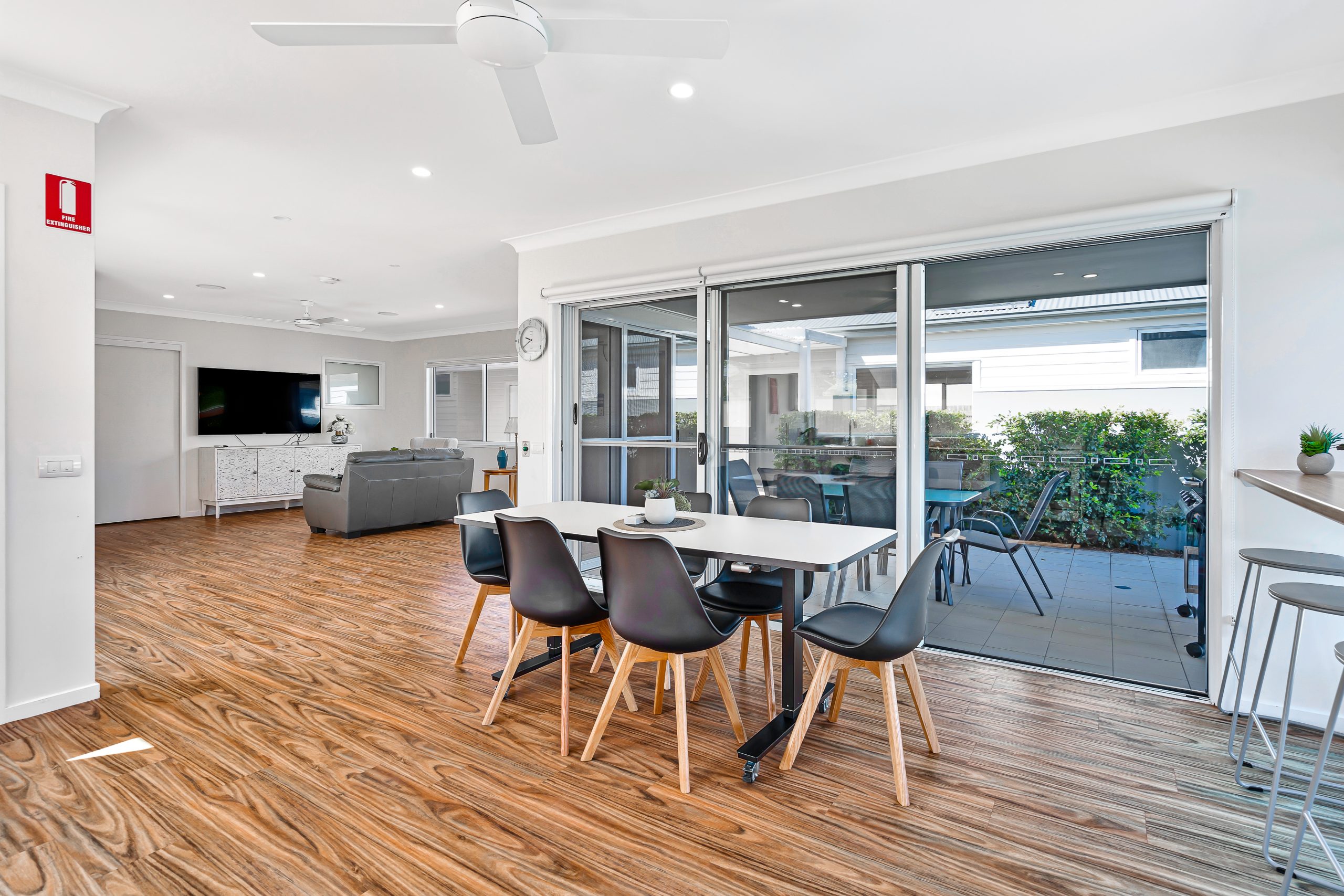 Modern open-plan dining and living area with wooden flooring, black dining chairs around a white table, large glass doors leading to a covered patio, and a ceiling fan above.