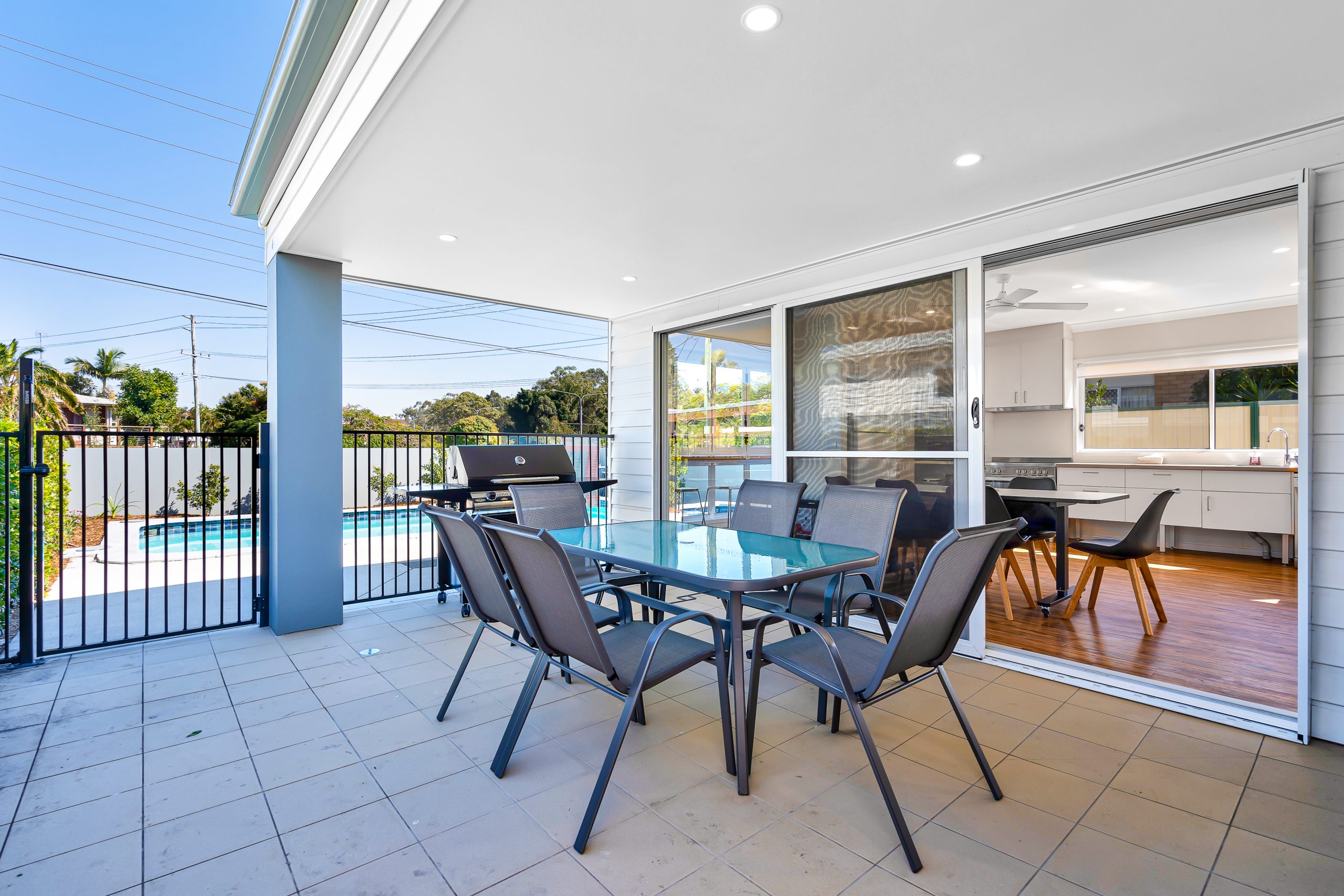 A modern outdoor patio with a glass dining table, six chairs, and a barbecue grill overlooks a fenced pool area. The space connects to a bright kitchen through sliding glass doors.