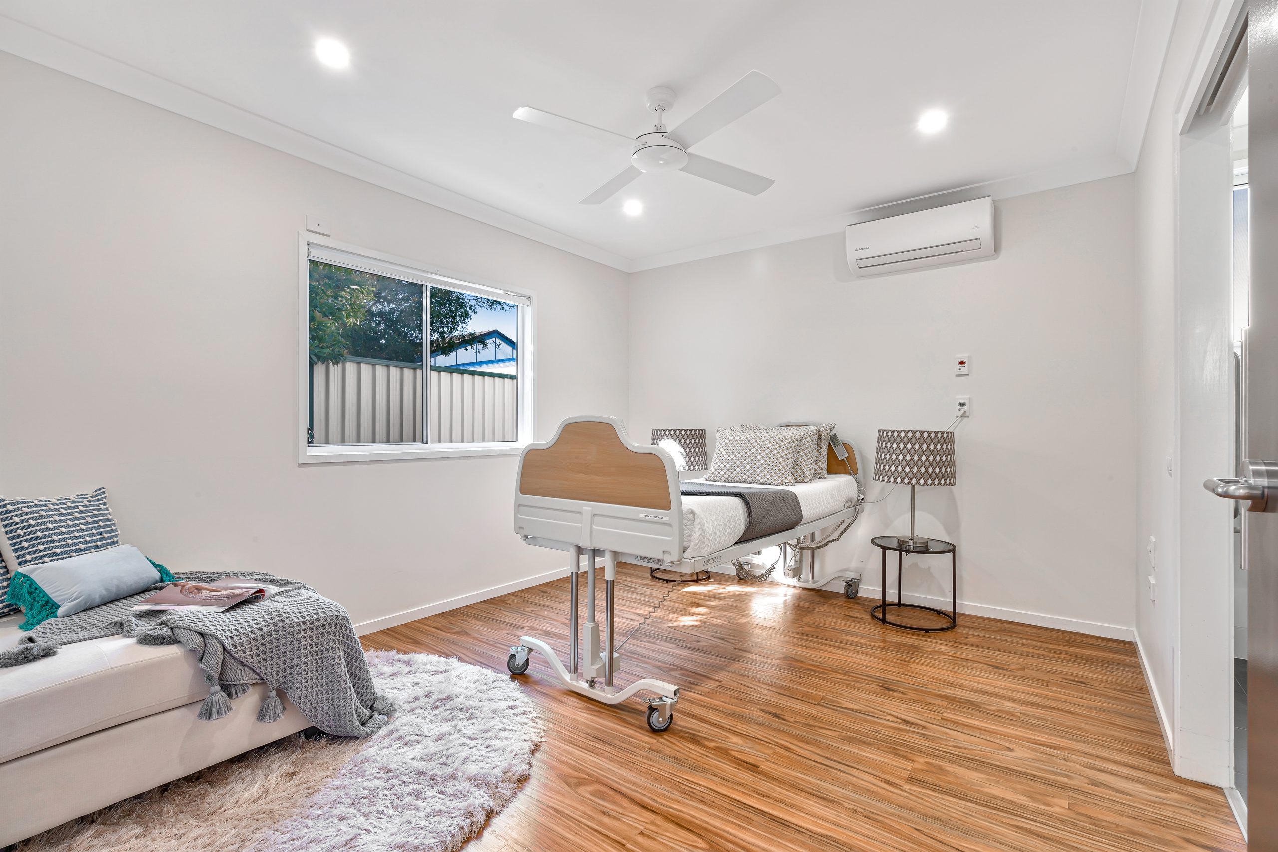 A clean, modern hospital room with a single adjustable bed, wooden floors, a window with outdoor view, a small sofa with blankets and pillows, a ceiling fan, and an air conditioning unit on the wall.
