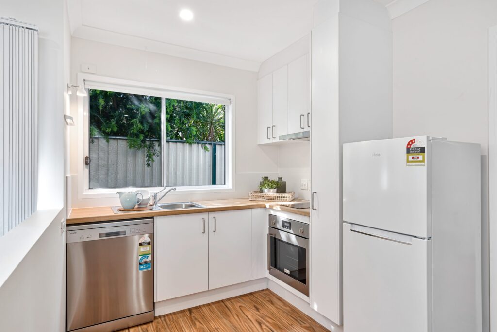 Modern kitchen with white cabinets, wood countertops, stainless steel dishwasher, built-in oven, white fridge, sink under a window, potted plants, and sunlight streaming in from outside greenery.