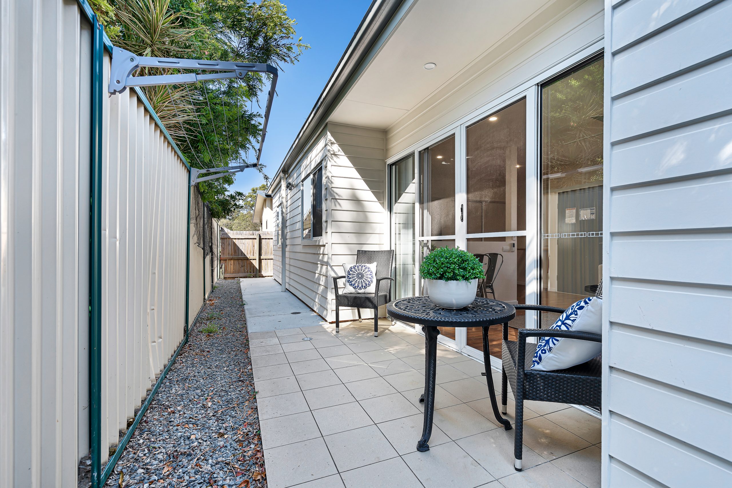 A small outdoor patio area with tiled flooring, a black round table, two chairs, and a potted plant. The area is beside a cream-colored fence and a house with large sliding glass doors.