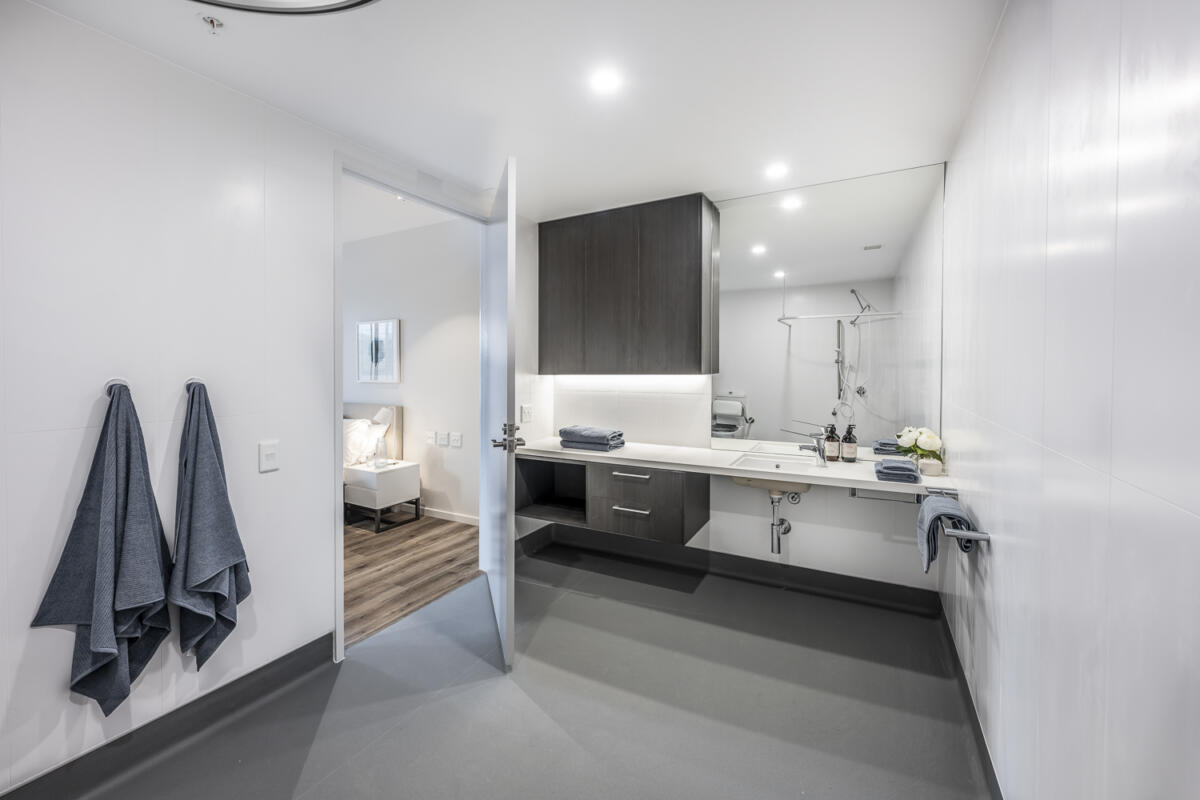 Modern, accessible bathroom with wide doorway, white walls, grey floor, large mirror, vanity with dark cabinets, and two navy towels hanging on the wall; bedroom visible through the open door.