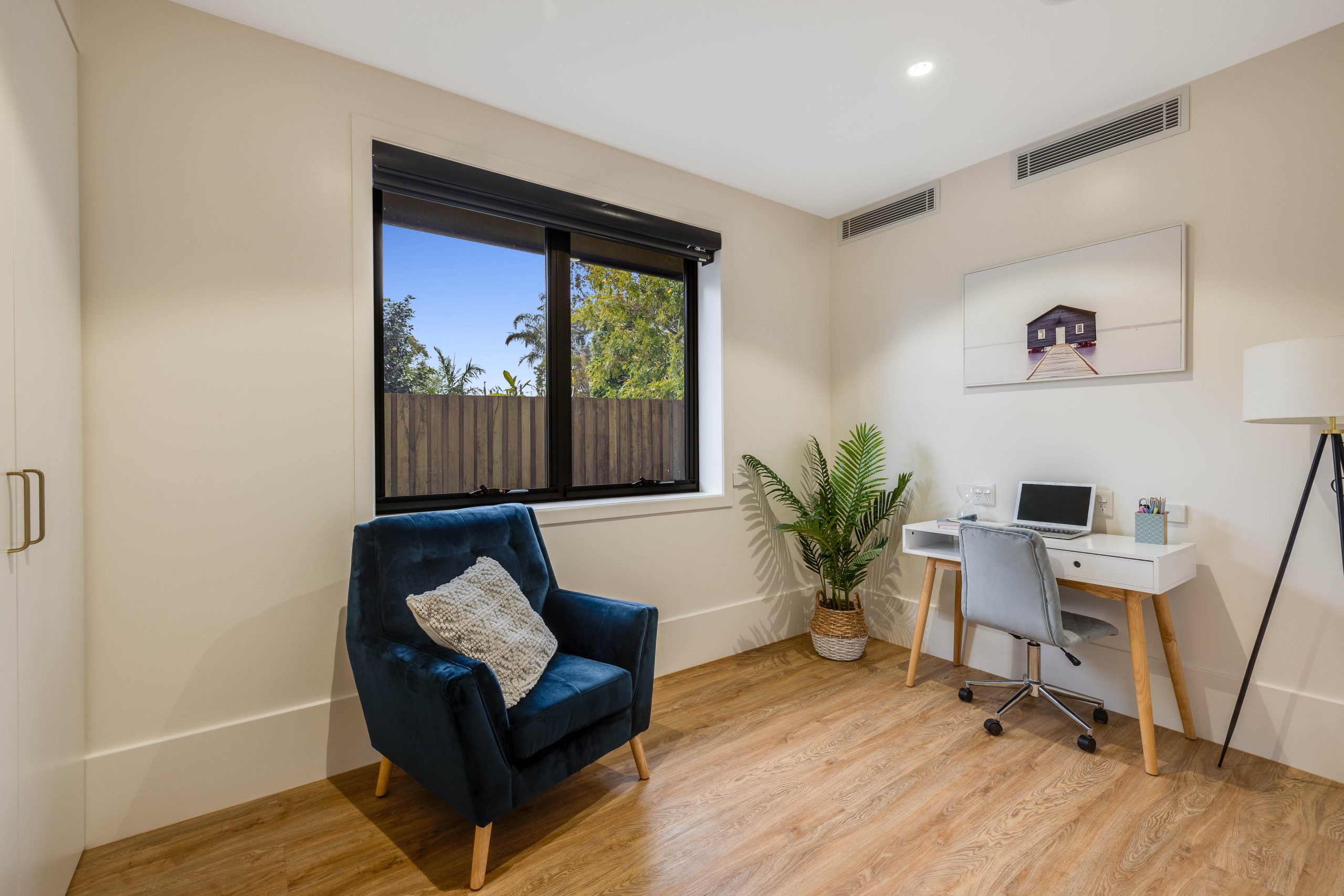 Modern home office with a blue armchair, decorative pillow, white desk with a laptop, potted plant, floor lamp, wall art, and a large window letting in natural light, with wood flooring and neutral walls.