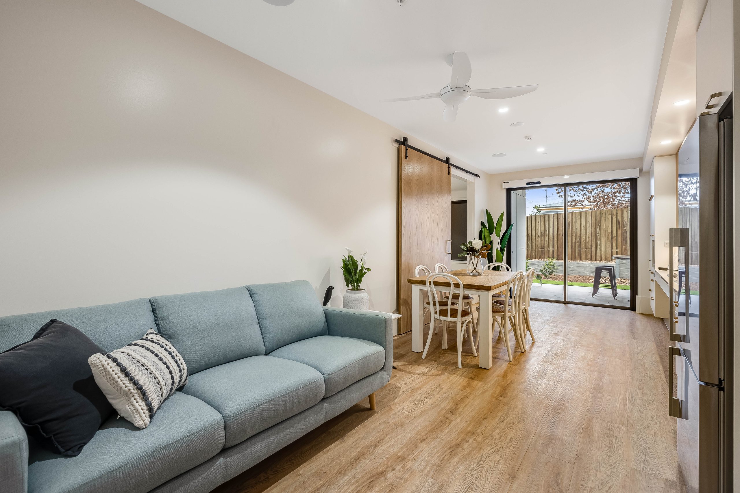 Modern living and dining area with a light blue sofa, wooden floors, dining table with white chairs, sliding barn door, potted plants, and large glass doors leading to a backyard with a wooden fence.
