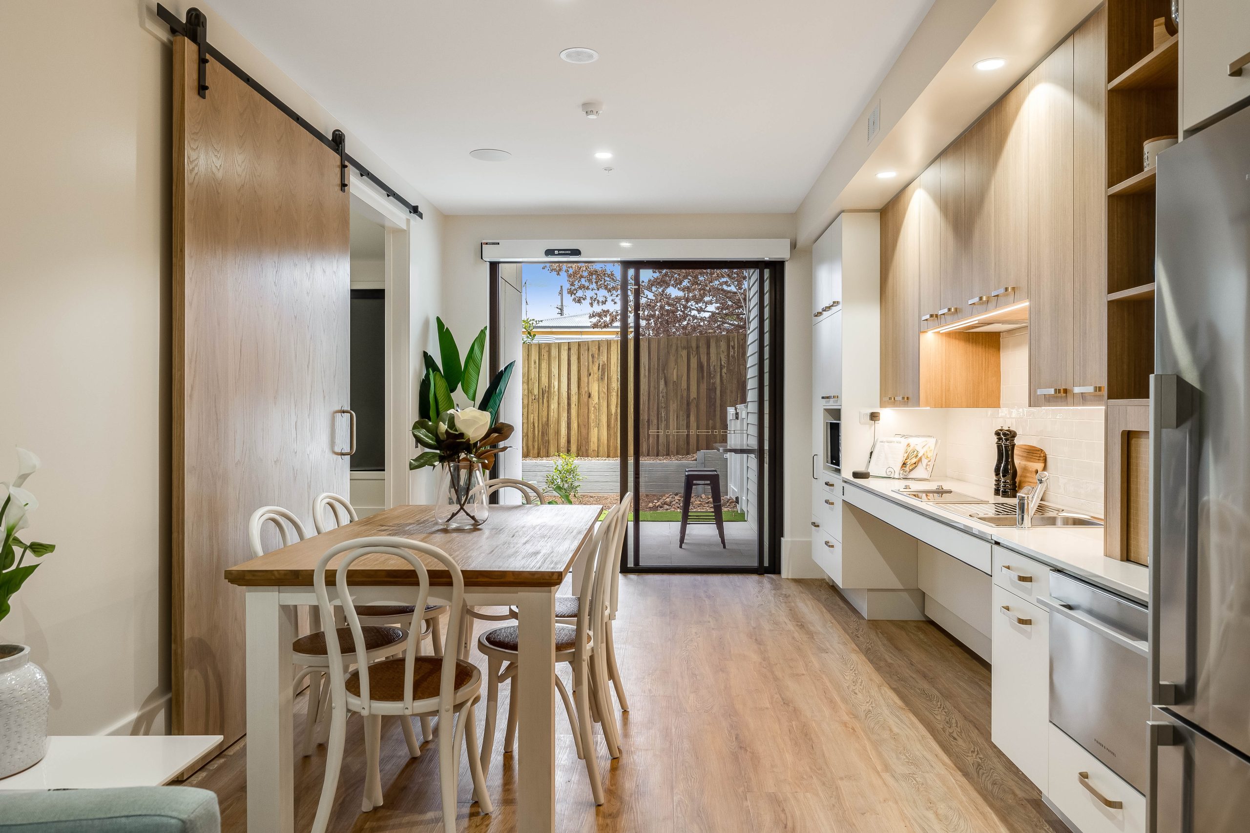 Modern kitchen and dining area with wood floors, a wooden dining table with four chairs, a sliding barn door, sleek white cabinetry, plants, and glass sliding doors leading to a small outdoor patio with a fence.