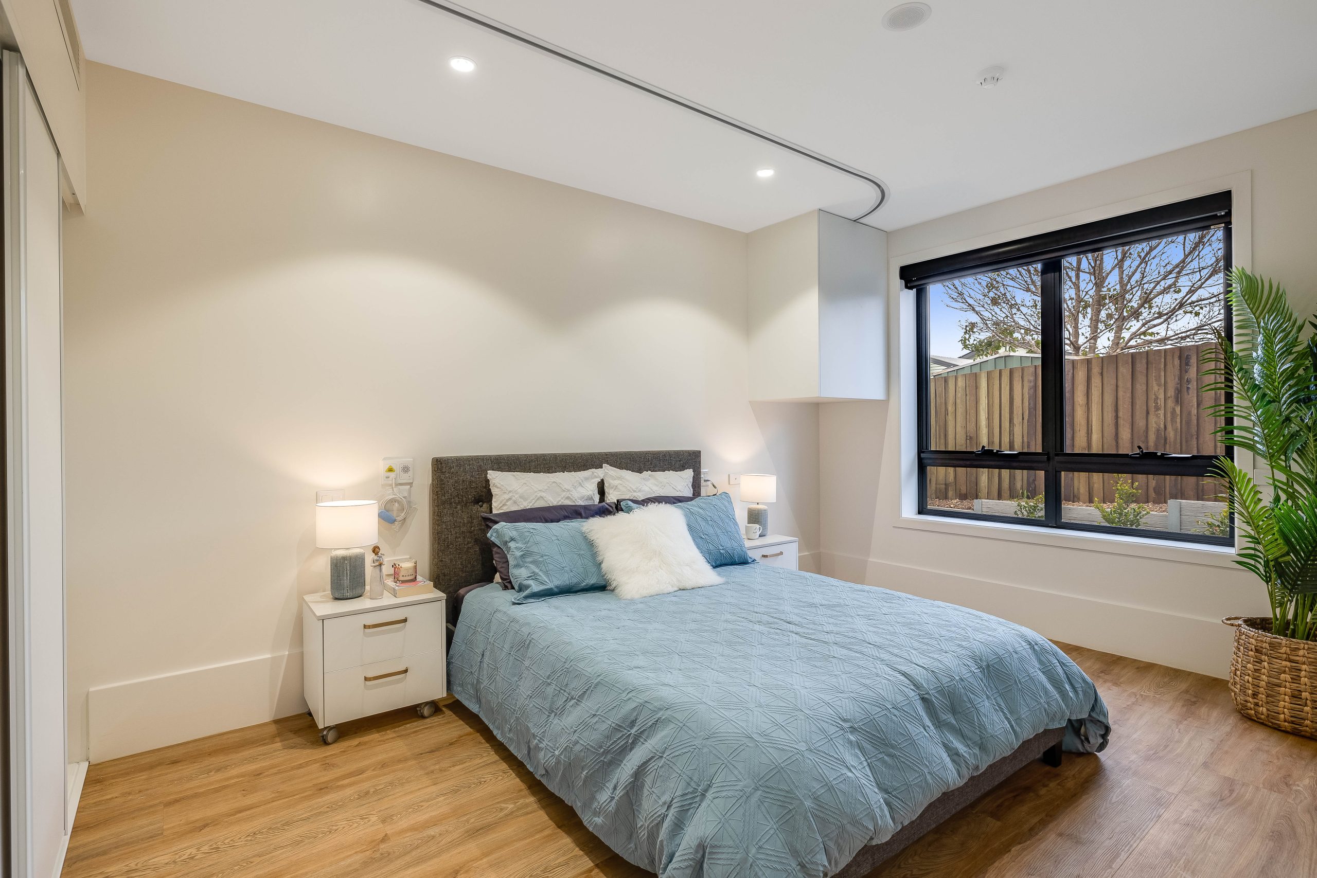 Modern bedroom with a double bed covered in a blue quilt, two white nightstands with lamps, a large window with black blinds, wooden floor, and a potted plant in the corner. Walls are light-colored and the room is tidy.
