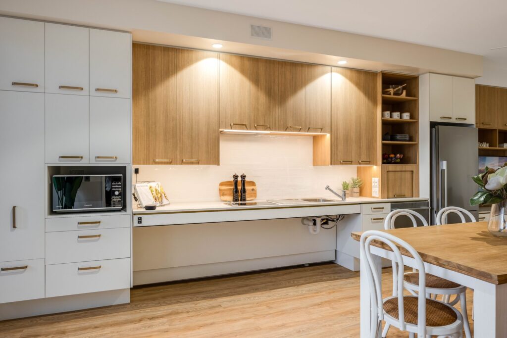 Modern kitchen with light wood cabinets, white countertops, built-in appliances, a sink, and utensils. A dining table with white chairs and a floral centerpiece is in the foreground. Warm lighting creates a cozy atmosphere.