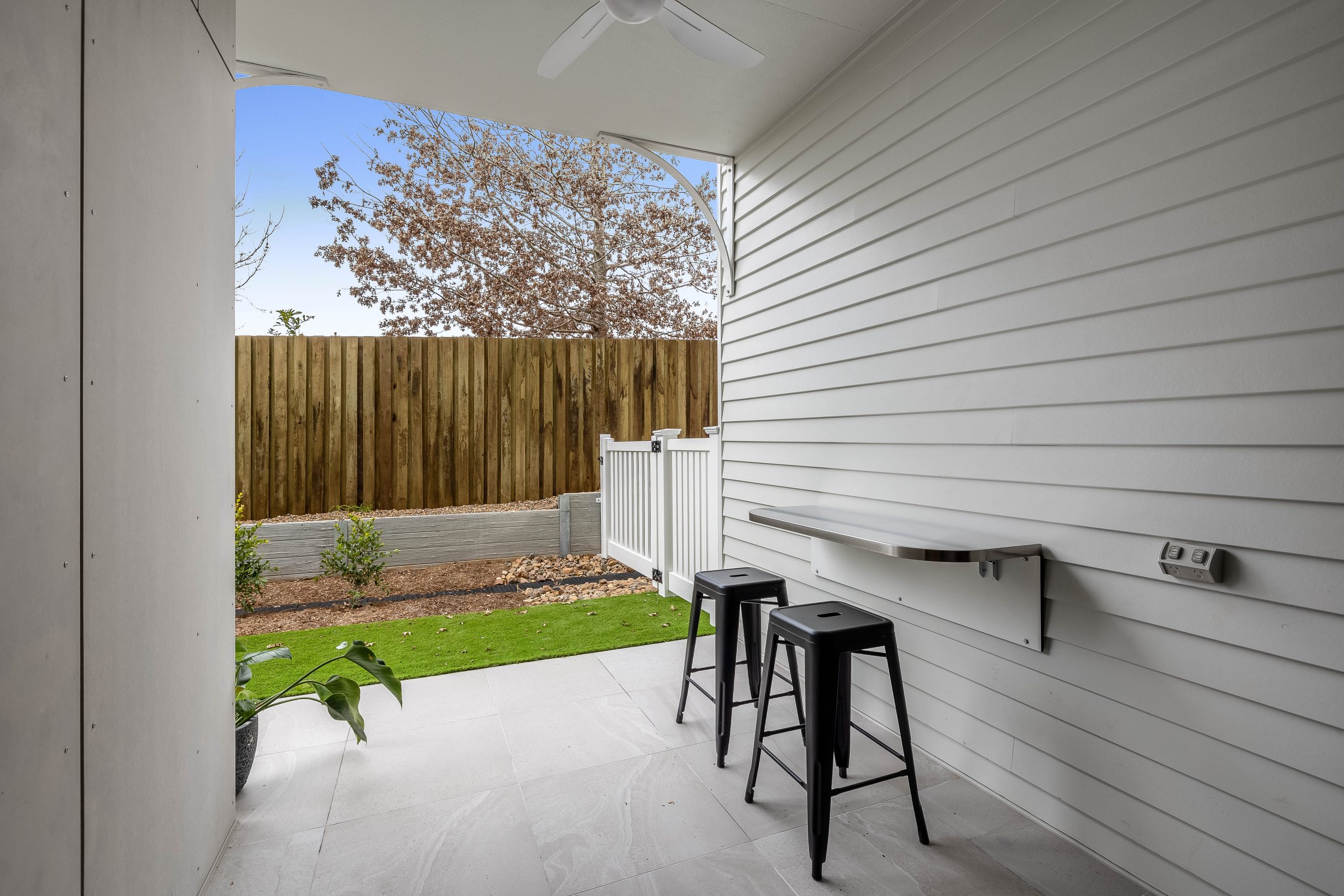 A small covered patio with white siding, a fold-down wall table, two black stools, ceiling fan, and view of a fenced backyard with green grass and planted shrubs.