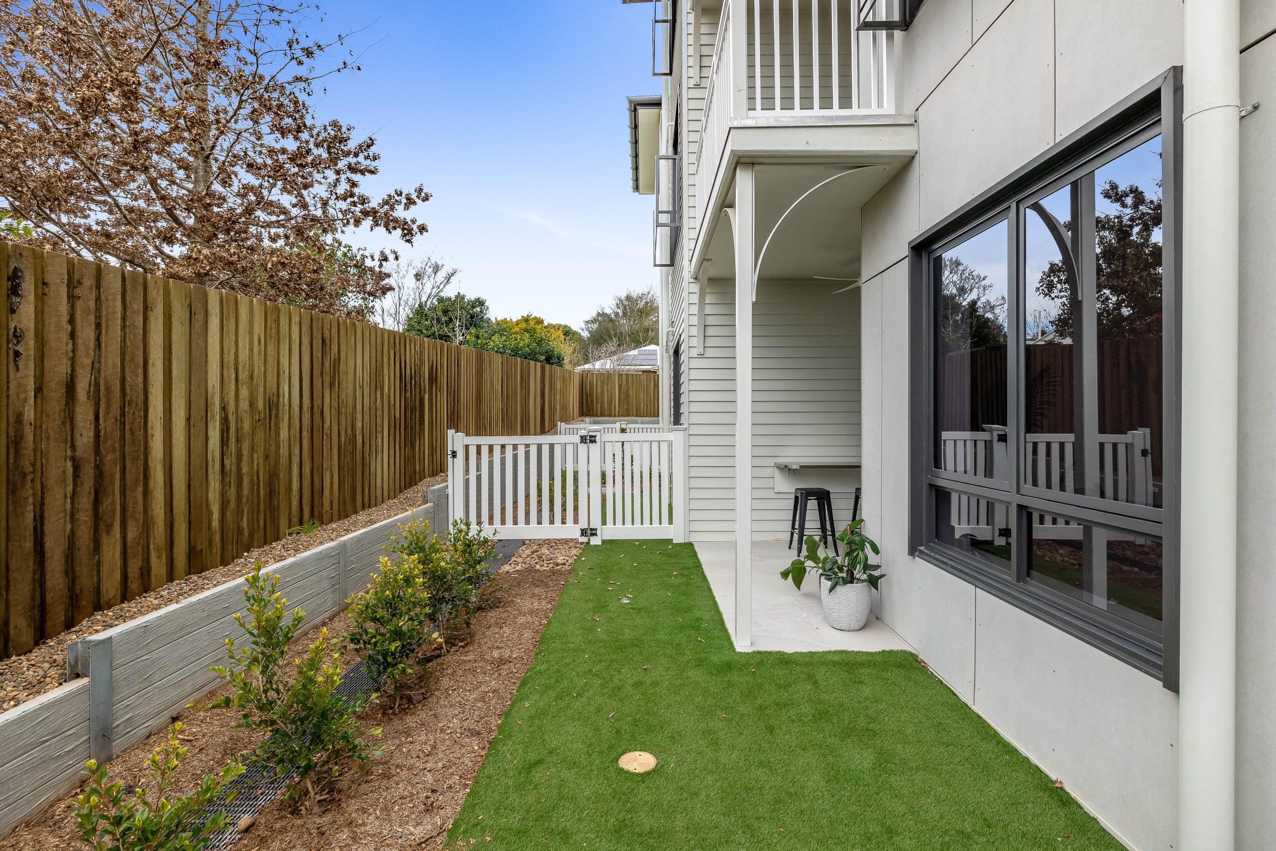 A narrow, modern backyard with artificial grass, a small garden bed along a wooden fence, a patio area with a stool and potted plant, and a white gate at the end.